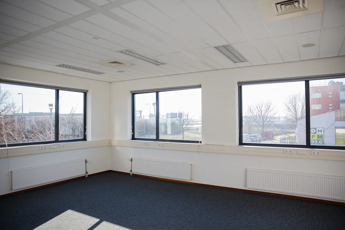 Leeg kantoor met tapijtvloer en grote ramen aan de Laan van 's-Gravenmade, met uitzicht op buitengebied en omliggende gebouwen.